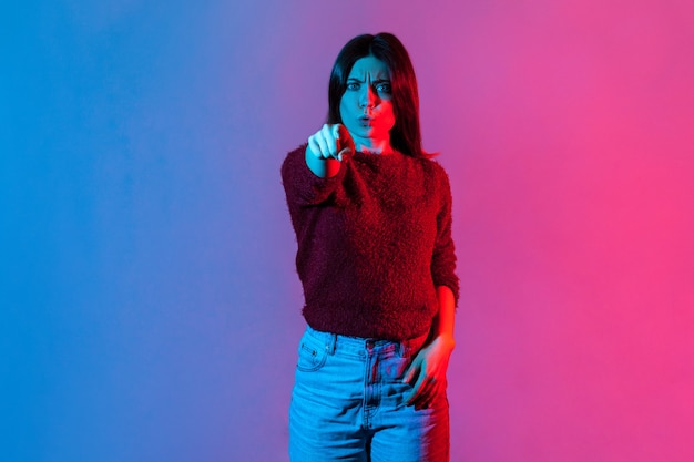 Neonlichtporträt einer strengen herrischen Frau, die unzufrieden wütend die Stirn runzelt und mit dem Finger auf die Kamera zeigt