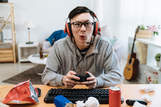 Nerd geek jovem adulto jogando no console de vídeo gosta de jogo online. jogador masculino asiático concentrado tendo competição no computador usando joystick no quarto bagunçado em casa. lixo de junk food dentro de casa.