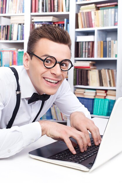 Nerd informático. Vista lateral del joven emocionado en camisa y pajarita escribiendo algo en la computadora y mirando a la cámara con una sonrisa