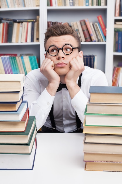 Nerd soñando. Joven aburrido en camisa y pajarita sentado en la mesa en la biblioteca y sosteniendo la cabeza entre las manos mientras dos pilas de libros en primer plano