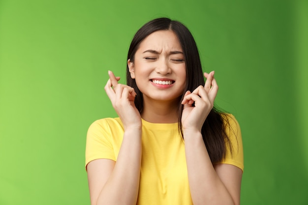 Nerviosa linda chica morena asiática ansiosa de ganar, preocuparse por los resultados importantes, cruzar los dedos, buena suerte, orar fortuna recibir promoción, apretar los dientes cerrar los ojos suplicando, soporte fondo verde