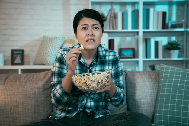 Nervöse asiatische zuschauerin, die nachts zu hause fernsieht und auf der couch im dunklen wohnzimmer sitzt. Junges Mädchen isst Schüssel Popcorn Junk Food und starrt auf Fernsehbildschirm mit Horror-Gruselfilm