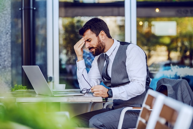 Foto nervoso caucasiano empresário barbudo bonito de terno sentado no café e segurando a cabeça. na mesa estão laptop, café e óculos. problemas no trabalho.