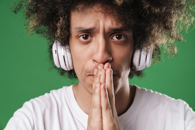 Foto nervoso jovem encaracolado posando isolado na parede verde ouvindo música com fones de ouvido, esperançoso, por favor gesticule.