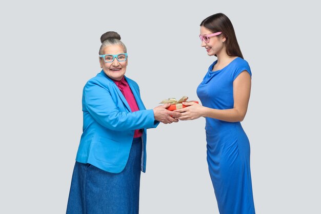 Neta de vestido azul sorri e dá um presente para sua adorável avó, olhando para a câmera com cara de surpresa. amizade compreensão mútua. indoor, studio shot, isolado em fundo cinza.