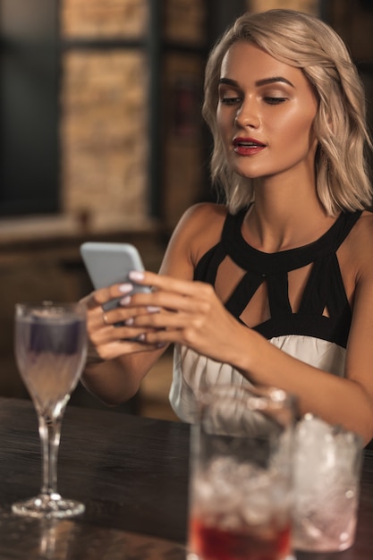 Únete a mi. Encantadora mujer rubia sentada en la barra del bar y enviando mensajes de texto a sus amigos, invitándolos a unirse a ella en el bar