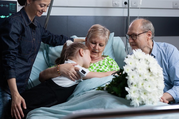 Netinha dando buquê de flores para a avó doente. Paciente idosa recebendo presente floral de parentes em visita hospitalar. Menina abraçando velha doente no quarto da clínica médica.