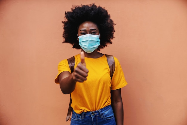 Nette afrikanische Frau, die draußen mit Maske auf ihrem Gesicht steht und Daumen oben zeigt. Covid19 Ausbruch.