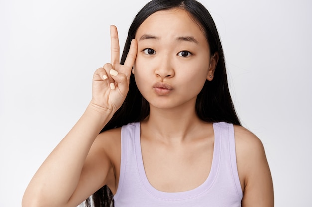 Nette asiatische Frau, die kawaii Friedenszeichen nahe sauberem Gesicht ohne Make-up zeigt, stehend auf Weiß. Platz kopieren