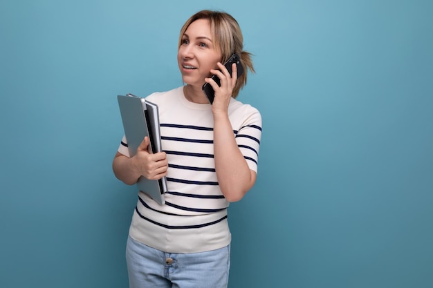 Nette blonde Frau, die am Telefon spricht und einen Laptop in ihren Händen auf blauem Hintergrund mit Kopie hält