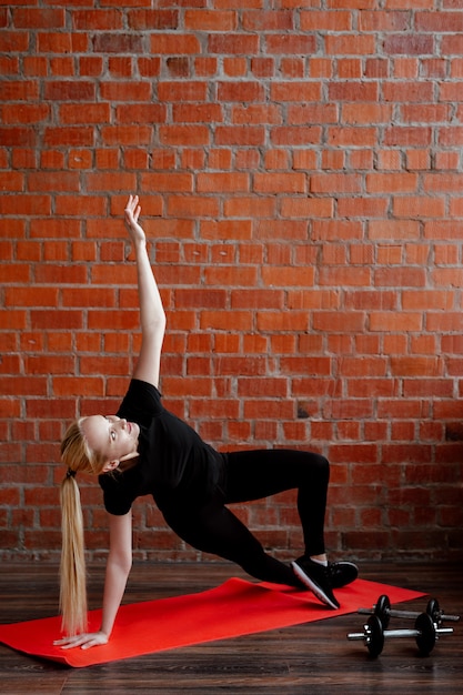 Nette blonde Frau, die Fitness auf dem Hintergrund einer Backsteinmauer des Hauses während der Quarantäne tut.