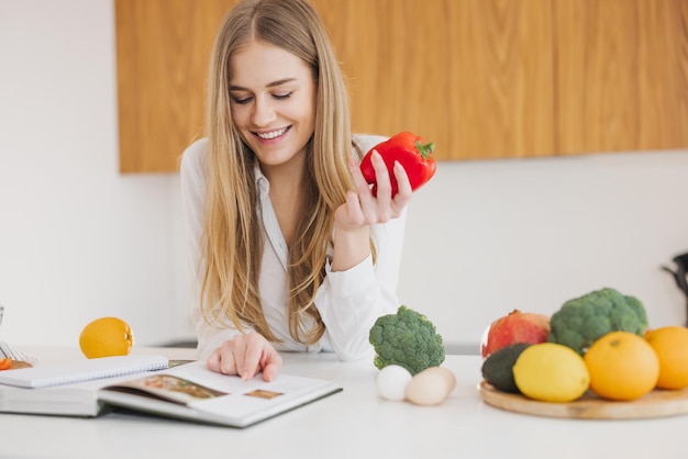 Nette blonde Frau, die Paprika hält und Rezeptbuch in der Küche auf dem Tisch zwischen Kochzutaten liest