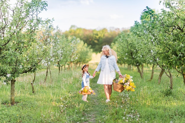 Nette blonde Frau mit ihrer kleinen Tochter in einem blühenden Frühlingsgarten Mutter und Tochter, die in der Natur mit Blumen in einem Korb spazieren gehen