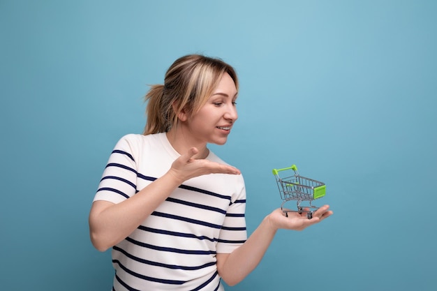 Nette blonde junge Mutter, die Einkaufswagenminiatur auf blauem Hintergrund mit Kopienraum zeigt