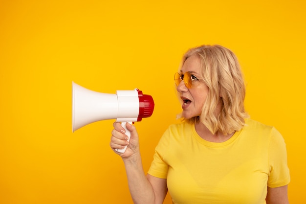 Nette blonde mittlere Frau sprechen im Megaphon, das über der gelben Wand isoliert wird.