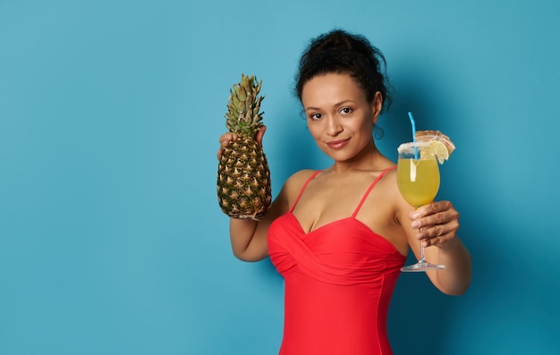 Nette Brünette im Badeanzug mit einer Ananas in der Hand, die einen leckeren Cocktail zur Kamera zeigt, isoliert