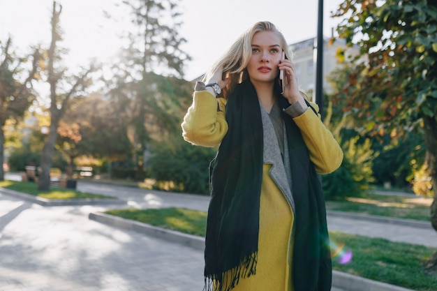 Nette emotionale attraktive blonde Frau im Mantel mit Smartphone geht die Stadtstraße. Kommunikation während des Spaziergangs, Lebensstil, Straße.