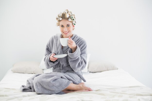 Nette entspannte Blondine in den Haarlockenwicklern Kaffee trinkend