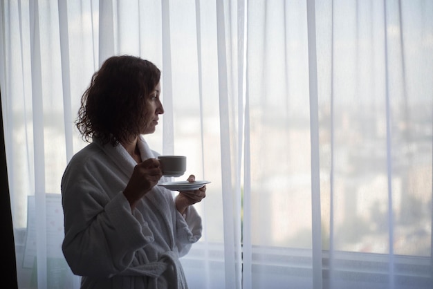 Nette Frau trinkt Kaffee und schaut durch das Fenster im Hotelzimmer