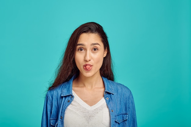 Nette Frau zeigt ihre Zunge, blaue Wand, positive Emotion. Gesichtsausdruck, weibliche Person, die im Studio vor der Kamera schaut, emotionales Konzept, Gefühle