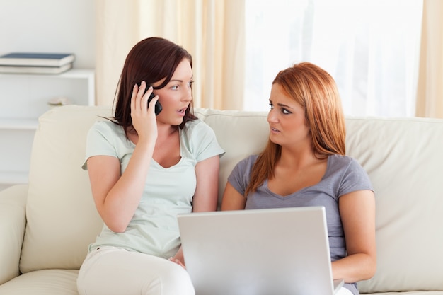 Nette Frauen, die auf einem Sofa mit einem Laptop und einem Telefon sitzen