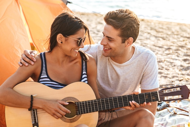 Nette Freunde, die Paar draußen am Strand lieben, sitzen, während auf der Gitarre spielen