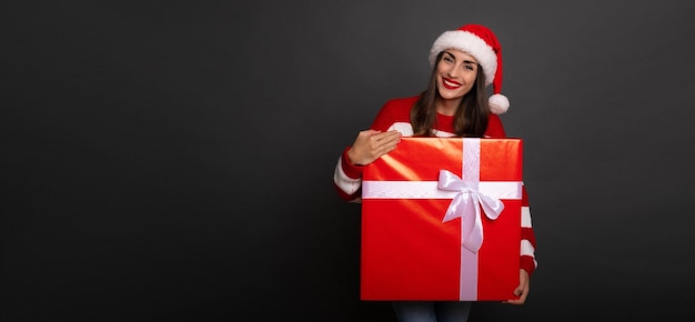 Nette glückliche junge brünette Frau in Weihnachtsmütze mit der großen roten Geschenkbox in den Händen hat Spaß beim posieren isoliert im Studio