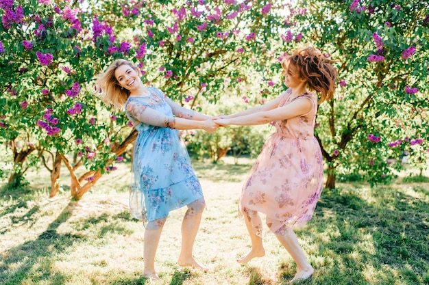 Nette glückliche Zwillingsschwestern in den schönen Kleidern, die im blühenden Sommerpark wirbeln.