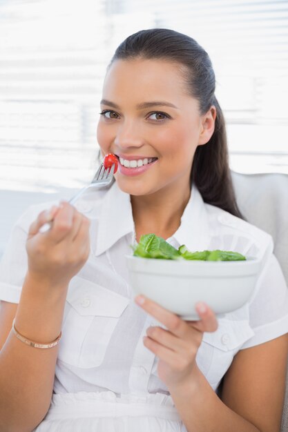Nette hübsche Frau, die den gesunden Salat sitzt auf Sofa isst