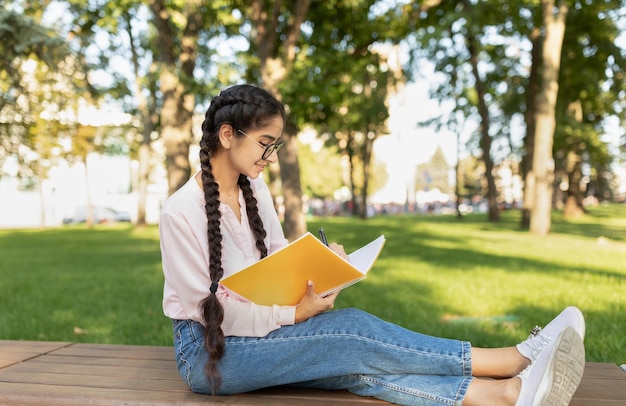 Nette indische Studentin, die draußen in Notizbuch schreibt und online lernt, während sie Freizeit verbringt