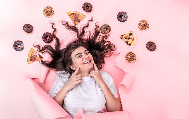 Foto nette junge brünette frau auf einem rosa papierhintergrund mit pizza und donuts, draufsicht, fast-food-konzept.