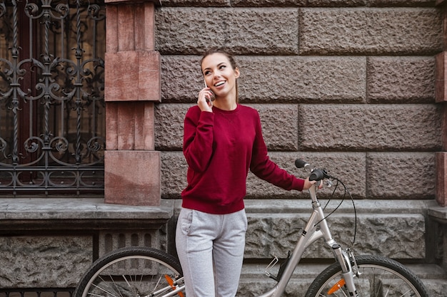 Nette junge Dame, die mit ihrem Fahrrad in der Stadt spazieren geht, während sie in der Nähe der Wand steht und mit ihrem Telefon spricht