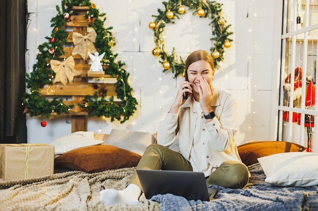 Nette junge Frau, die am Telefon spricht, um sich mit Laptop und Technologie auf Weihnachten vorzubereiten, während sie zu Hause im Urlaub auf dem Bett neben dem Weihnachtsbaum sitzt