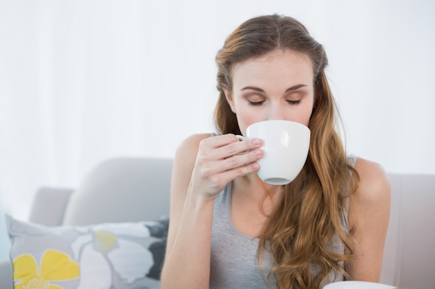 Nette junge Frau, die auf dem Sofa trinkt von der Schale sitzt