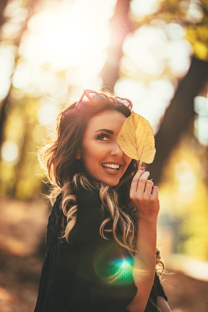 Nette junge Frau, die im sonnigen Wald in Herbstfarben genießt. Sie hält goldenes Blatt und schaut auf die Kamera.
