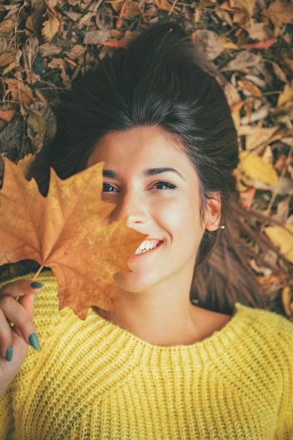 Nette junge Frau, die im sonnigen Wald in Herbstfarben genießt. Sie liegt auf der Herbstwiese und blickt hinter Blättern in die Kamera.