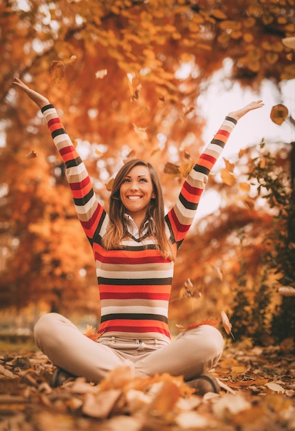 Nette junge Frau, die im sonnigen Wald in Herbstfarben genießt. Sie sitzt auf dem mit Blättern bedeckten Boden und hat Spaß.