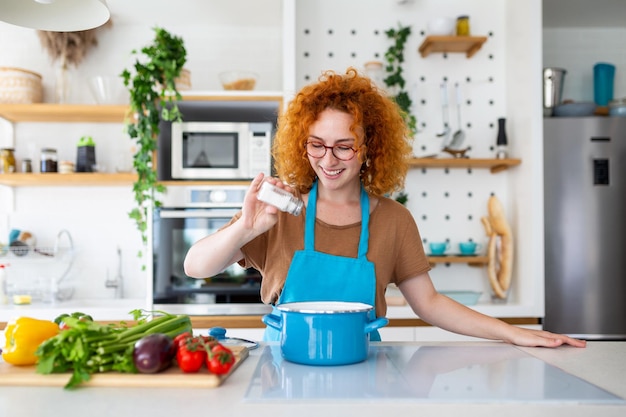 Nette junge Frau, die kocht und der Mahlzeit Würze hinzufügt, lacht und Zeit in der Küche verbringt