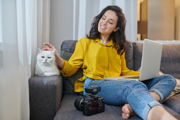 Nette junge Frau, die mit ihrer Katze neben ihr auf dem Sofa sitzt