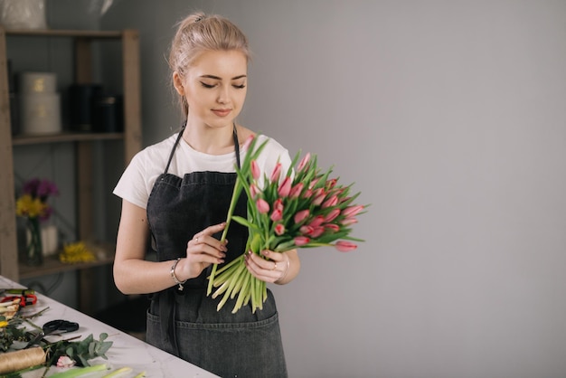 Nette junge Frau Floristin mit Schürze, die Blumenarrangements aus frischer Tulpe am Tisch auf weißem Hintergrund macht. Konzept der Arbeit mit Blumen, Blumengeschäft.