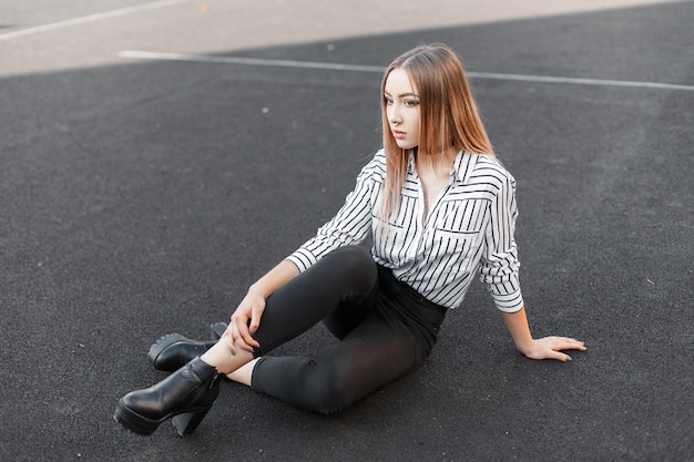 Nette junge Frau in einem stilvollen schwarz-weiß gestreiften Hemd in schwarzen Jeans in Lederschuhen mit einer durchbohrten Nase sitzt auf dem Stadion an einem sonnigen warmen Sommertag.