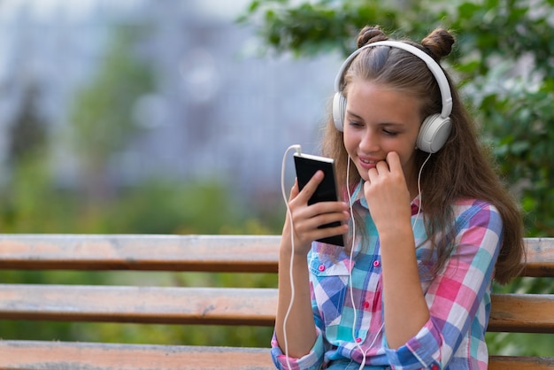 Nette junge Frau mit einem süßen Lächeln, das draußen auf einer Bank sitzt und ihre Musik auf Kopfhörern hört