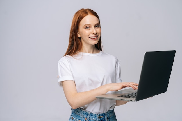 Nette junge Frau mit T-Shirt und Jeanshose, die Laptop-Computer hält und in die Kamera schaut