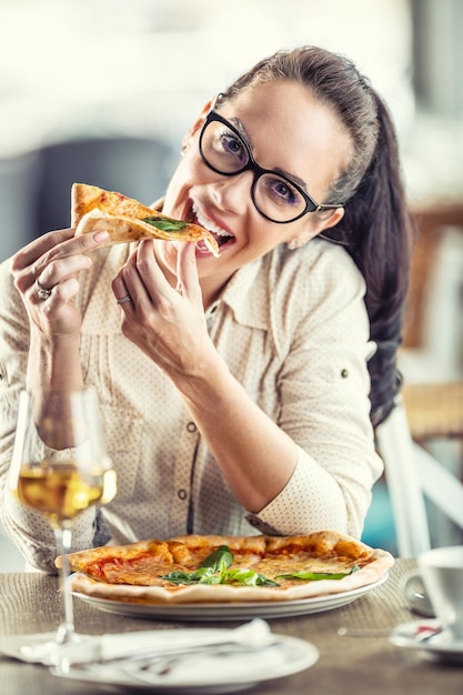 Nette junge Frau neigt ihren Kopf, während sie in ein Stück Pizza beißt