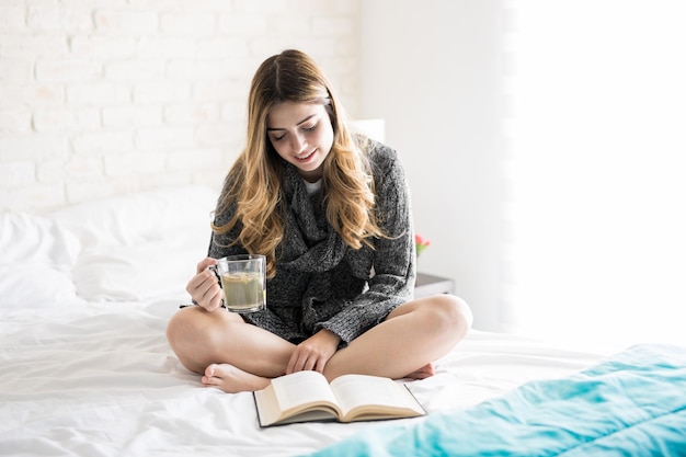 Nette junge gemütliche Frau, die auf ihrem Bett sitzt, während sie ein Buch liest und Tee trinkt