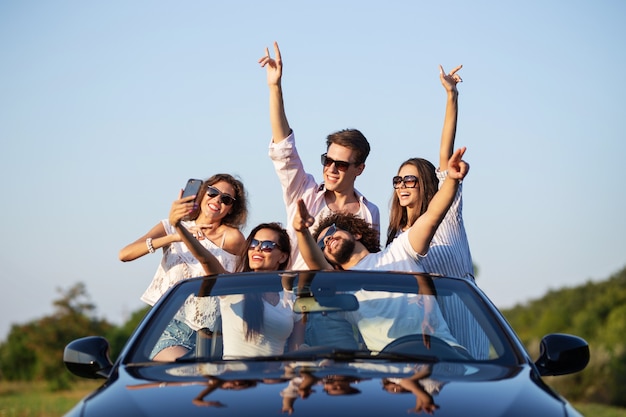 Nette junge Mädchen und Jungs mit Sonnenbrille sitzen in einem schwarzen Cabrio auf der Straße, halten ihre Hände hoch und machen an einem sonnigen Tag Selfies. .