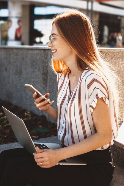 Nette junge rothaarige Frau mit Sommersprossen, die draußen auf der Treppe lachend ein Smartphone in der Hand einen Laptop auf ihren Beinen lachend lacht.