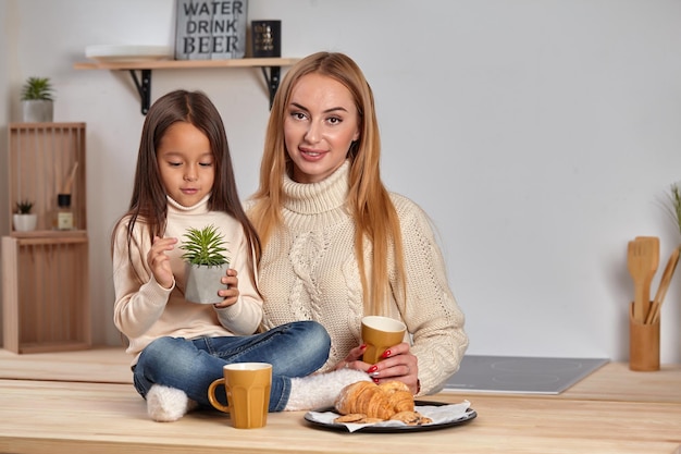 Nette lächelnde Mutter, die ihrem Kind, das auf der Küche sitzt, ein Croissant gibt