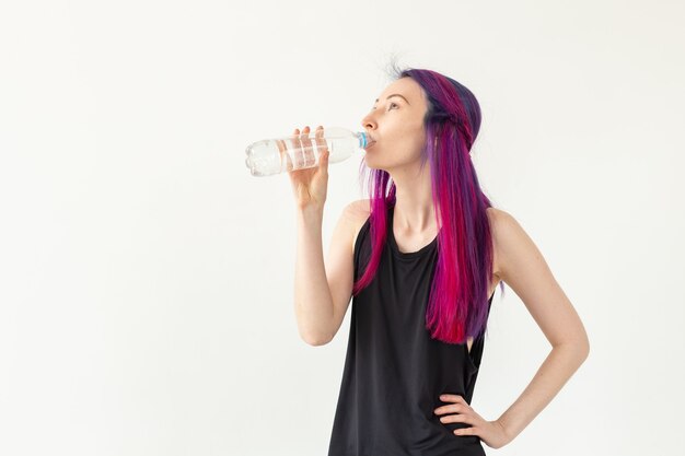 Nette schlanke junge asiatische Frau mit gefärbtem Haartrinkwasser nach dem Training im Fitnessstudio. Konzept der Wiederauffüllung des Wasserhaushalts und eines gesunden Lebensstils. Weiße Wand mit Kopierraum