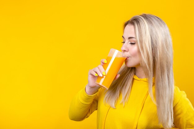 Nette schöne junge blonde frau in der gelben lässigen sportlichen kleidung, die orangensaft vom glas trinkt,